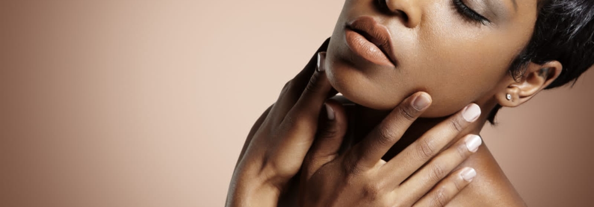 Une femme aux cheveux courts pose, les yeux fermés, sur un fond marron clair. Ses mains effleurent délicatement son cou et ses épaules, et elle paraît sereine et détendue. Dakar