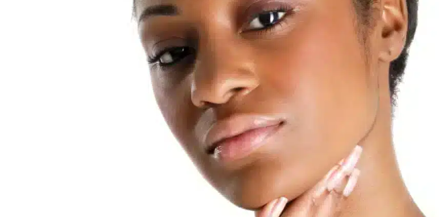 Une femme aux cheveux courts se touche délicatement le menton de la main droite, s'interrogeant sur son hyperpigmentation. Son expression est neutre, son regard légèrement hors champ sur un fond blanc. Dakar