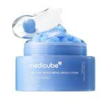 Un pot bleu de Medicube Hyaluronic Moisturizing Capsule Cream est ouvert, révélant des capsules bleues et blanches. Une spatule transparente avec une capsule repose sur le couvercle, rappelant la Prime+ Studio Base Réductrice de Pores d'Essence Cosmetics. Dakar