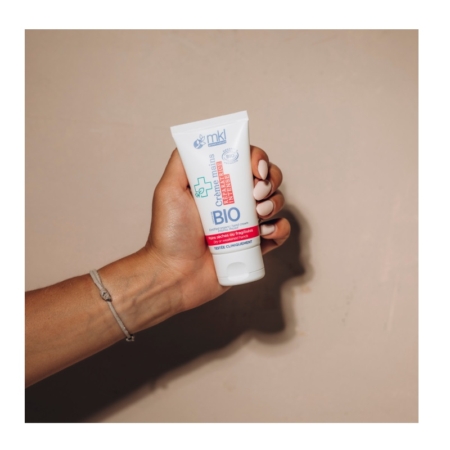 Une main aux ongles peints en blanc tient un tube de MKL Crème mains certifiée BIO Réparatrice - 50ml sur un fond beige uni. Un fin bracelet est porté au poignet. Dakar