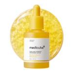 Un flacon compte-gouttes jaune étiqueté "Medicube Sérum à l'Acide Kojique au Curcuma et à la Niacinamide Teint Irregulier 30ml" se trouve devant un fond jaune rond et texturé, suggérant un sérum avec des particules en suspension. Dakar