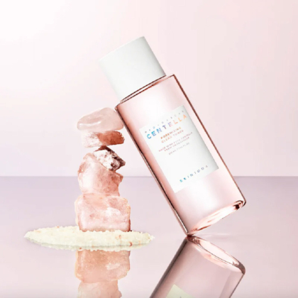 Un flacon rose de SKIN1004 Madagascar Centella Tonique Fraîcheeur Anti Pores Dilatés et Matifiant Peaux Mixtes à Grasses 210 ml s'appuie sur des cristaux roses sur une surface rose pastel, avec des granules blancs dispersés à la base. Dakar