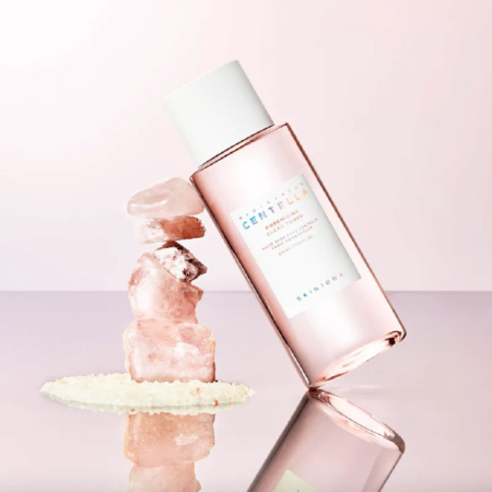 Un flacon rose de SKIN1004 Madagascar Centella Tonique Fraîcheeur Anti Pores Dilatés et Matifiant Peaux Mixtes à Grasses 210 ml s'appuie sur des cristaux roses sur une surface rose pastel, avec des granules blancs dispersés à la base. Dakar