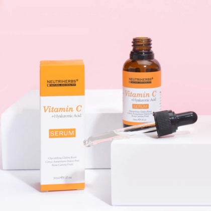 Un flacon orange et blanc étiqueté « NEUTRIHERBS Vitamine C Sérum 30 ml » est placé à côté de sa boîte correspondante sur une surface blanche sur fond rose. Un compte-gouttes contenant du sérum se trouve devant le flacon. Dakar