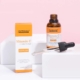 Un flacon orange et blanc étiqueté « NEUTRIHERBS Vitamine C Sérum 30 ml » est placé à côté de sa boîte correspondante sur une surface blanche sur fond rose. Un compte-gouttes contenant du sérum se trouve devant le flacon. Dakar