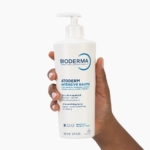 Une main tient un flacon-pompe blanc de Bioderma Atoderm Intensive Baume à côté de Bioderma ATODERM Huile De Douche 1L. Le baume apaise les peaux très sèches et irritées ; le flacon de 500 ml a un texte bleu et noir. Dakar