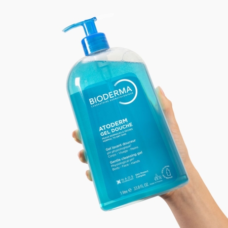 Une main tient un flacon de 1 litre de Bioderma ATODERM Gel Douche Nettoyant Douceur, un gel nettoyant doux pour le corps et les mains, doté d'une pompe bleue et d'un emballage transparent. Dakar