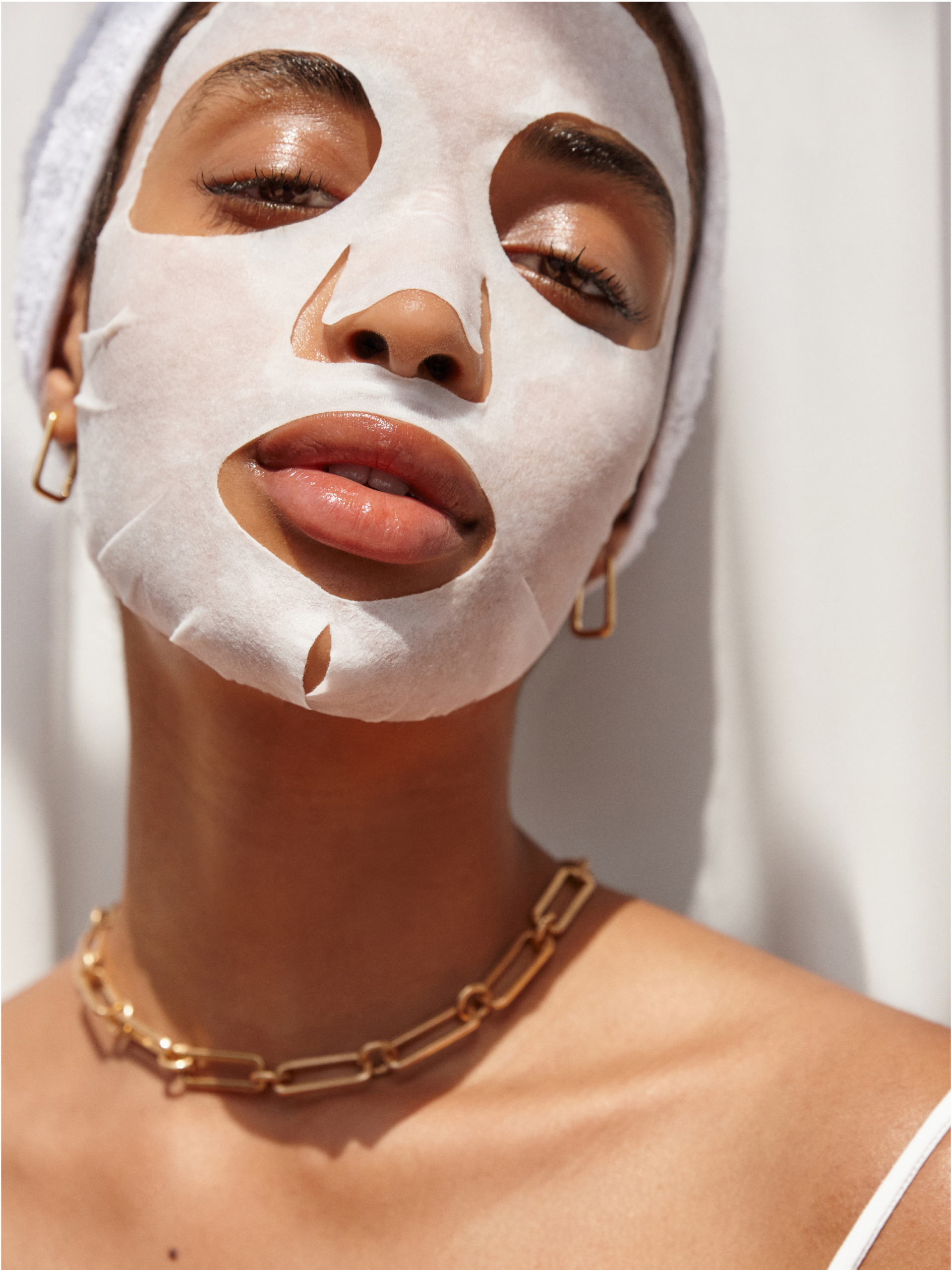 Une femme porte le masque en tissu « Skin Booster Illuminateur » de Byphasse (1 pièce), un collier à chaîne dorée et des boucles d'oreilles créoles. Une serviette enroulée autour de la tête, elle fait face à l'objectif avec assurance, sur un fond clair. Dakar