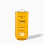 Un flacon jaune de « Shampooing à la Kératine CHEVEUX SECS ET ABÎMÉS » de Byphasse, d'une contenance de 500 ml, destiné aux cheveux secs et abîmés, muni d'une pompe blanche et portant une étiquette mettant en avant l'éclat et la nutrition, projetant une ombre sur un fond blanc. Dakar