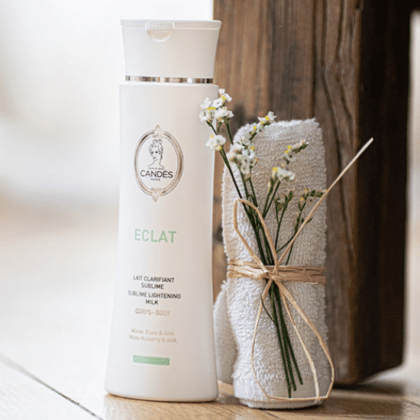 Une bouteille de Candés Lait Clarifiant Sublime 400 ml d'Univers Cosmetix trône à côté d'une serviette blanche roulée nouée avec de la ficelle et de petites fleurs blanches