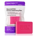 Un savon rose Carbon Theory "Savon Anti Imperfections à la Niacinamide et de l'Acide Citrique 100g" et sa pochette violette et blanche affichent les détails du produit, les ingrédients et la marque sur le savon et l'emballage. Dakar