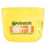 Un pot jaune de crème sorbet Garnier Vitamine C Fresh & Bright Glow Booster avec un graphique de tranches de citron et un texte sur la vitamine C se trouve à côté du brillant à lèvres Juicy Bomb Shiny Lipgloss 01 Proud Pitaya 10ml d'Essence Cosmetics, formant ainsi un duo de beauté radieux. Dakar