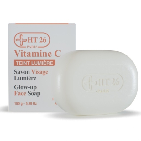 Une barre blanche de Ht26 Savon Visage Lumière Vitamine C Illuminateur 150g, estampillée "HT 26 Paris", se trouve devant sa boîte blanche étiquetée avec un texte orange et gris : "Ht26 Vitamine C Teint Lumière Glow-up Face Soap. Dakar