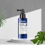 Un flacon pompe bleu de L'Oréal Serioxyl Denser Hair Sérum Traitement Activateur de Densité 90ml repose sur une plateforme en pierre blanche, entourée de feuilles tropicales vertes et sur un fond texturé gris. Dakar