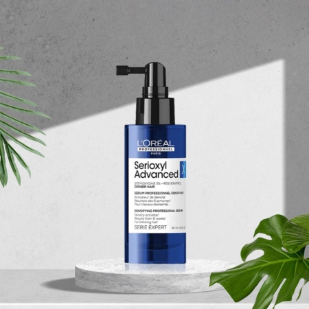 Un flacon pompe bleu de L'Oréal Serioxyl Denser Hair Sérum Traitement Activateur de Densité 90ml repose sur une plateforme en pierre blanche, entourée de feuilles tropicales vertes et sur un fond texturé gris. Dakar