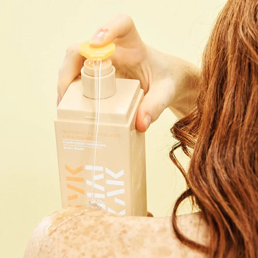 Une personne aux cheveux mouillés utilise une pompe jaune pour faire couler sur son épaule un nettoyant pour le corps contenu dans un flacon beige. L'étiquette, comme celle de DODIE Biberon Tétine Plate 0-6 Mois Mer Plastique 270 ml, met en avant les acides glycolique et lactique pour un nettoyage en douceur. Dakar