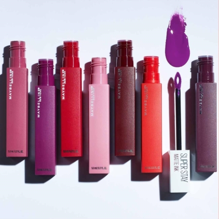 Sept tubes de Maybelline Rouge à lèvres New York Superstay Ink sont disposés dans différentes teintes, avec un tube ouvert montrant son applicateur et un échantillon violet au-dessus. Dakar