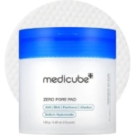 Un récipient blanc avec un couvercle bleu étiqueté "Medicube Zero Pore Pad 2.0 Tampons Exfoliant Anti Pore Dilate et Acné 70 pads" se trouve devant un tampon rond texturé, mettant en évidence l'AHA, le BHA, le Panthénol, l'Allantoïne et l'Hyaluronate de sodium. Dakar