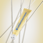 Un tube jaune de Nineless Ampoule Capillaire à la Caféine Root Biome 20ml avec un embout pointu est suspendu parmi des tiges métalliques, une goutte à l'extrémité de l'applicateur, sur un fond dégradé beige et jaune. Dakar