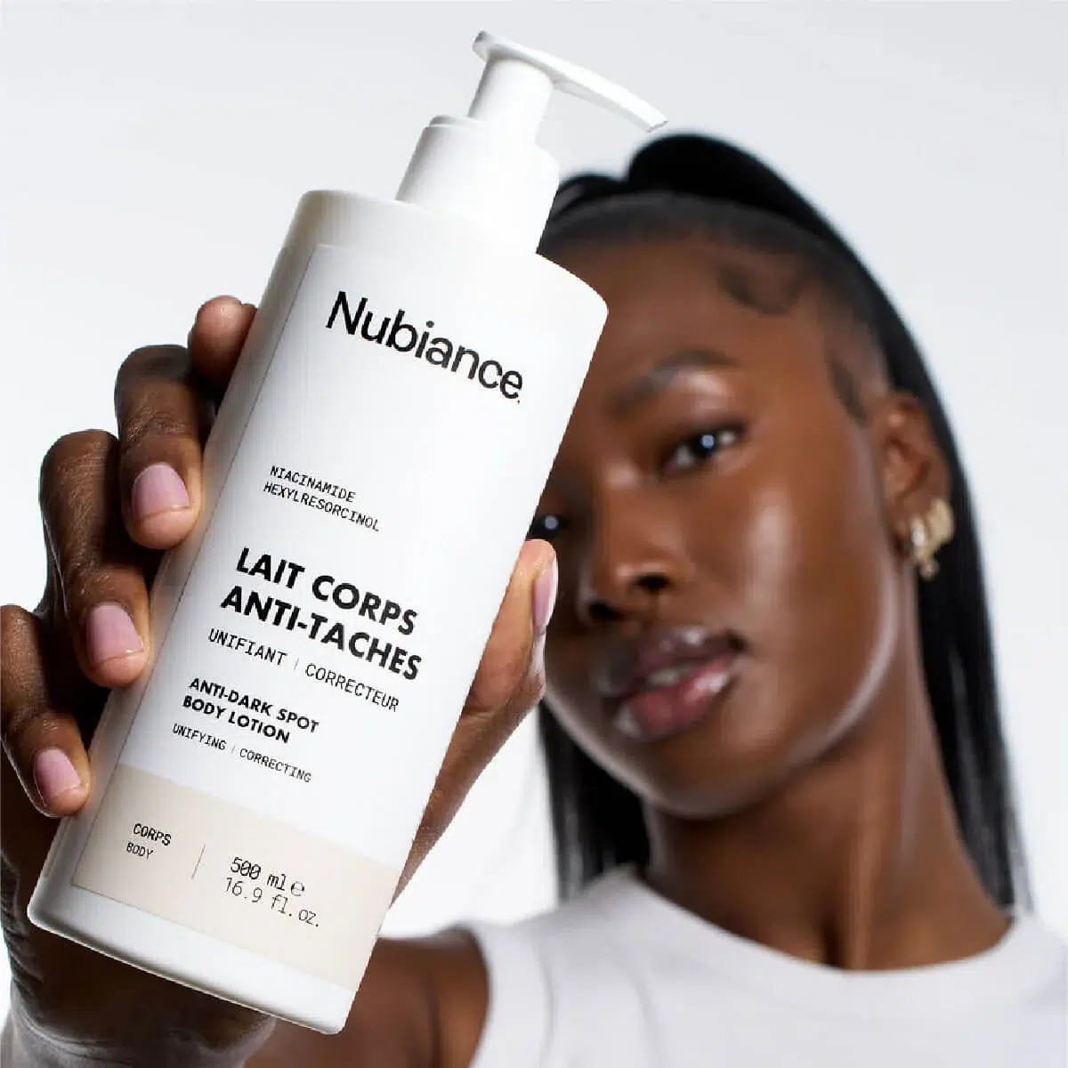 Une femme à la peau foncée tient un flacon de Nubiance Lait Corps Unifiant & Hydratant HRB-3 500ml, dont l'étiquette est clairement visible sur un fond blanc uni. Dakar