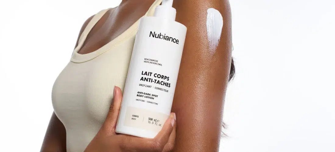 Une personne vêtue d'un débardeur beige tient un flacon de Nubiance Lait Corps Unifiant & Hydratant HRB-3 500ml, avec une trace de lotion appliquée sur le haut du bras. Dakar