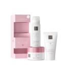 Un coffret cadeau pour femme Rituals The Ritual Of Sakura rose et blanc avec gel douche moussant, gommage corporel et crème pour le corps est présenté à côté d'une boîte ouverte sur le thème des fleurs de cerisier roses. Dakar