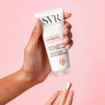 Une main tient un tube de SVR Cicavit+ Crème Apaisante Réparation Accélérée Anti-Marques 40ml sur un fond rose, tandis qu'une autre applique la crème avec une noisette sur le bout du doigt, prête pour une application en douceur. Dakar