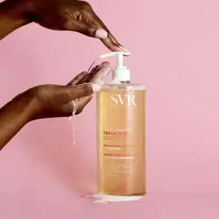 Les mains pressent la pompe de SVR TOPIALYSE Huile Lavante Anti-Irritation Relipidante pour peaux sensibles, libérant l'huile nettoyante dans les paumes ouvertes. Le flacon transparent avec pompe et inscription blanches se détache sur un fond rose tandis que l'huile coule doucement en cascade en dessous. Dakar
