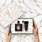 Une femme tient un coffret cadeau parfum femme Yves Saint Laurent BLACK OPIUM EDP COFFRET ouvert sur une surface en marbre, révélant le parfum, le spray de voyage et la lotion pour le corps dans un élégant emballage blanc avec des fissures dorées et des logos YSL. Dakar