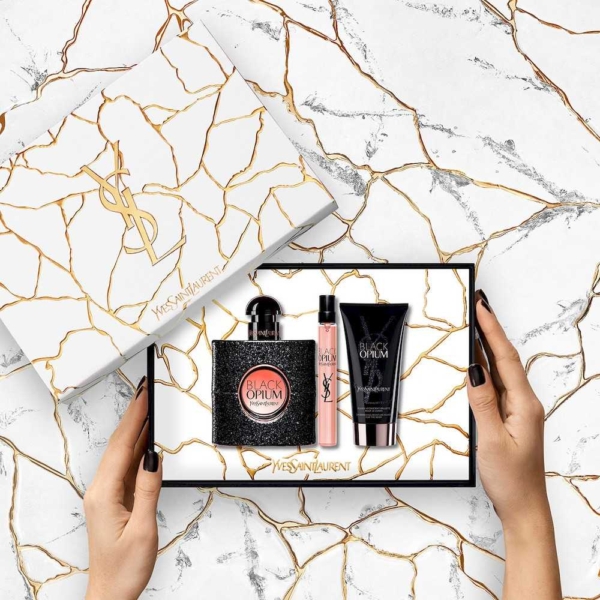 Une femme tient un coffret cadeau parfum femme Yves Saint Laurent BLACK OPIUM EDP COFFRET ouvert sur une surface en marbre, révélant le parfum, le spray de voyage et la lotion pour le corps dans un élégant emballage blanc avec des fissures dorées et des logos YSL. Dakar
