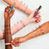 L'image présente trois bras aux tons de peau différents, chacun orné d'échantillons de brillant à lèvres Brillant à Lèvres Repulpant Extrême What The Fake d'Essence Cosmetics dans les teintes 01, 02 et 03. Chaque individu tient un tube correspondant de brillant à lèvres. Les teintes varient du nude au rose et au marron. Dakar