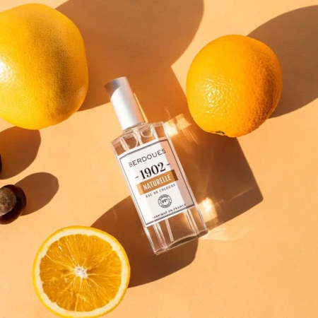 Un flacon d'Eau de Cologne BERDOUES Naturelle 125 ml repose sur une surface pêche, entouré d'agrumes frais - un citron entier, une orange entière et une orange tranchée - projetant des ombres douces et incarnant son essence. Dakar