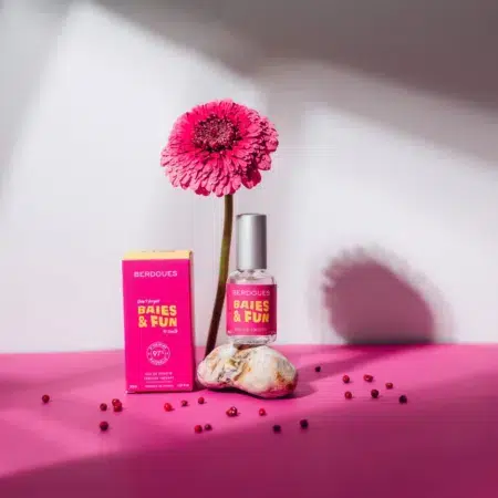 Un coffret de parfum rose étiqueté « BERDOUES Baies and Fun Eau de Toilette 30ml » côtoie un flacon assorti avec un gerbera rose en arrière-plan. Ils se détachent sur un fond rose et blanc, parsemé de perles rouges. Dakar