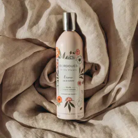 Un flacon d'eau de toilette Berdoues Mille Fleurs l'eau est posé sur un tissu texturé beige. Le flacon est rose avec des motifs floraux et un bouchon argenté, et il porte le texte "94% d'origine naturelle" indiquant les ingrédients naturels. Dakar