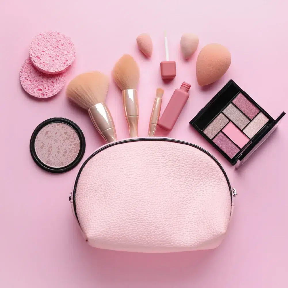 Une trousse de maquillage rose entourée de produits cosmétiques et d'outils sur fond rose. Comprend des éponges de maquillage, un poudrier, trois pinceaux, un rouge à lèvres et une palette de fards à paupières aux teintes neutres. Dakar