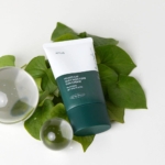 Un tube d'Anua Crème Solaire Spf50 Hydratante ET Soyeuse Heartleaf 50 ml avec SPF 50+ est posé sur des feuilles vertes brillantes, accompagnées de deux boules en verre transparent, créant une esthétique minimaliste et naturelle. Dakar