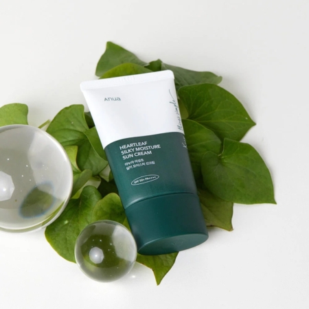 Un tube d'Anua Crème Solaire Spf50 Hydratante ET Soyeuse Heartleaf 50 ml avec SPF 50+ est posé sur des feuilles vertes brillantes, accompagnées de deux boules en verre transparent, créant une esthétique minimaliste et naturelle. Dakar