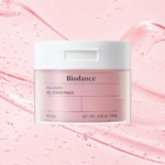 Un élégant pot blanc avec une étiquette rose, contenant 140 g de gel tonique anti-âge Biodance Collagène, se dresse sur un fond rose à texture liquide. Ce luxueux produit de soin contient 60 tampons pour un rituel de soins. Dakar