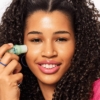 Une femme aux cheveux bouclés sourit en appliquant le Primer Stick Anti Cernes Jelly Grip 4,6 g d'Essence Cosmetics sur sa joue. Son rouge à lèvres rose et ses grandes boucles d'oreilles créoles accentuent son regard radieux. Le produit est présenté dans un petit contenant vert. Dakar
