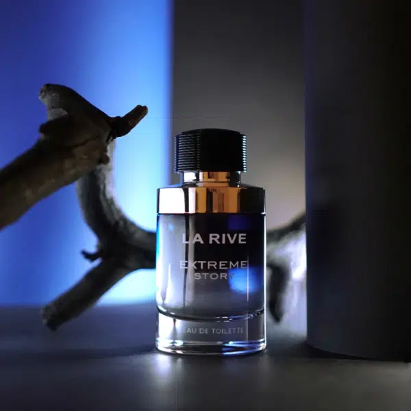 Un flacon d'eau de toilette « La Rive Extreme Story » repose sur une surface sombre, éclairée par une lueur bleue. Le flacon cylindrique a un bouchon argenté et une branche floue apparaît à l'arrière-plan. La scène est sombre et dramatique. Dakar
