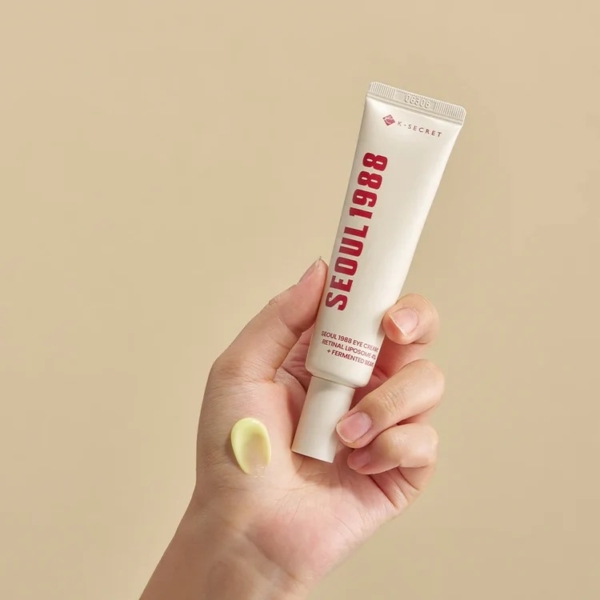 Une main tient un tube blanc de KSECRET Crème Contour Yeux SEOUL 1988 Liposome Rétinien 4% + Haricot Fermenté 30ml sur un fond beige neutre, en appliquant une noisette sur le dos de la main. Dakar