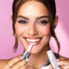 Une femme sourit tout en appliquant du brillant à lèvres à partir d'un tube. Elle a les cheveux bruns coiffés en deux mèches lâches et porte du maquillage pour les yeux. Ses ongles sont peints en argent, assortis à son haut. Le fond est rose. Dakar