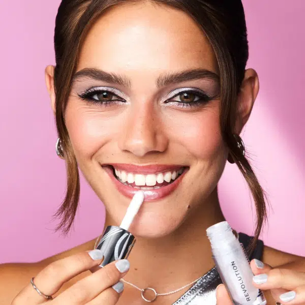 Une femme sourit tout en appliquant du brillant à lèvres à partir d'un tube. Elle a les cheveux bruns coiffés en deux mèches lâches et porte du maquillage pour les yeux. Ses ongles sont peints en argent, assortis à son haut. Le fond est rose. Dakar