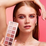 Une personne aux longs cheveux roux et aux boucles d'oreilles créoles tient une Palette de Fards à Paupières 12 Ombres de 8,4 g de Makeup Revolution près de son visage. Un fard à paupières rose pâle et un rouge à lèvres s'harmonisent avec le fond rosé, tandis qu'un fin collier en or orne son cou alors qu'elle pose élégamment avec une main sur la tête. Dakar