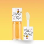 Tube de gloss à lèvres étiqueté « Honey & Milk » avec des illustrations d'abeilles et de bouteilles de lait sur un fond jaune dégradé. Le gloss est partiellement ouvert, laissant apparaître l'applicateur. Dakar