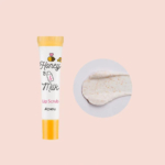 À gauche, un tube de gommage pour les lèvres A'pieu Miel et Lait, avec un bouchon jaune. À droite, une goutte de gommage, à la texture crémeuse et aux fines particules exfoliantes, est visible. Le fond est rose clair. Dakar
