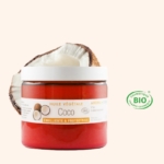 Un pot d'Aroma Zone Huile de Coco BIO 100ml avec un couvercle blanc et une étiquette française est placé devant une demi-coco, avec un logo cosmétique bio "BIO" vert à droite, sur un fond beige clair. Dakar