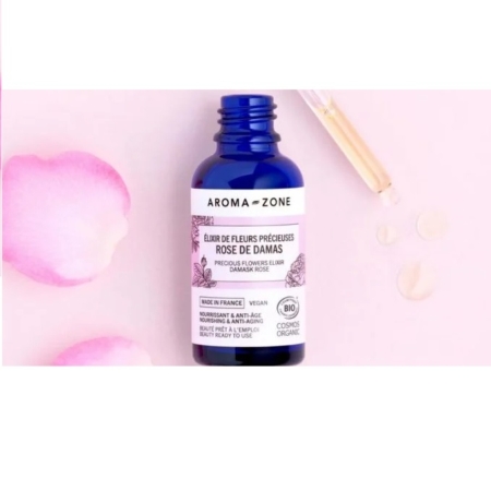 Un flacon bleu d'« Aroma Zone Élixir de fleurs précieuses de Rose de Damas BIO 30 ml » repose sur une surface rose ornée de pétales de rose, rappelant l'élégance du Sérum TOPICREM AH3. Un compte-gouttes distribue le liquide à côté du flacon. L'étiquette indique « Fabriqué en France », « Vegan » et « Bio ». Dakar