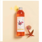 Un flacon d'Huile de Ricin BIO Aroma Zone de 100 ml repose sur une surface beige clair, partiellement renversé à côté d'une gousse de ricin rouge et verte et d'une feuille. Son étiquette porte l'inscription « Huile Végétale Ricin », rappelant subtilement l'Huile de Ricin Waam par ses détails raffinés. Dakar