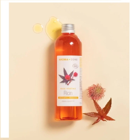 Un flacon d'Huile de Ricin BIO Aroma Zone de 100 ml repose sur une surface beige clair, partiellement renversé à côté d'une gousse de ricin rouge et verte et d'une feuille. Son étiquette porte l'inscription « Huile Végétale Ricin », rappelant subtilement l'Huile de Ricin Waam par ses détails raffinés. Dakar